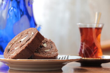 Chocolate banana roll cake slices on the plate with fork. Glass with black traditional turkish brewed tea in small glass and blue glass vase in background. Tea Time at home concept.