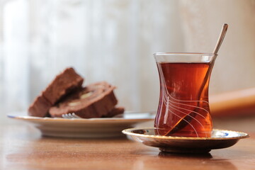 Glass with black traditional turkish brewed tea stands on an old wooden table. Chocolate banana roll cake in background. Turkish tea in traditional small glass. Hot drink. Tea Time at home concept