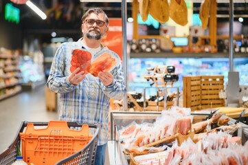 Elderly man buyer of meat department of grocery store, holding at meat sausage. Male pensioner customer orders meat snacks