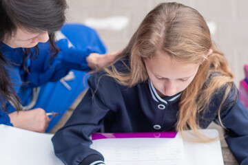 Naklejka premium smart girl solving math problems on her own while her mother sits nearby at wooden table on children's playground and watches,education concept,going back to school, girl in beautiful blue uniform