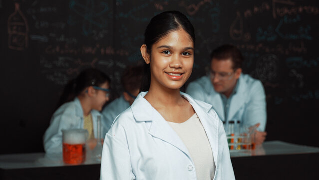 Girl looking at camera with arm folded while people doing experiment at laboratory. Cute student standing blackboard with chemical theory with blurring background at STEM science class. Edification.