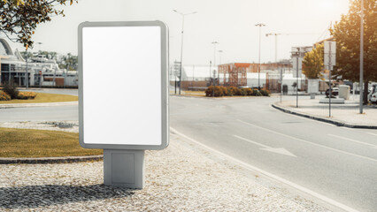 A bright mockup of a vertical outdoor empty billboard in an urban area. The sign stands on a stone-paved sidewalk by an empty street, with a blank white screen for advertisements