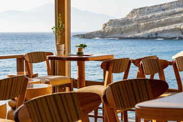 Empty restaurant tables and chairs directly on the beach, in the background the sea and a big rock, in the sun, Greece, Crete island, telephoto