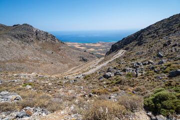 Serpentine road in barren rocky hillside with sea in distance in sunshine with clear blue sky,...