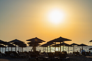 Silhouette of umbrellas and sunbeds on seaside beach with yellow sky, sun shining in front, backlit, telephoto