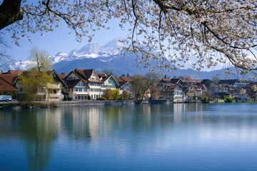 Fototapeta premium Cherry blossoms frame charming houses reflecting in the serene lake, with majestic snow-capped peaks visible in the distance. Generative AI