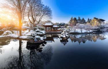 Obraz premium A wooden gazebo sits on the edge of a calm pond, surrounded by snow-covered trees and a clear blue sky.