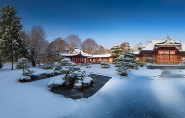 Fototapeta premium A traditional Japanese-style building complex with a red roof sits amidst a snowy, serene landscape with trees and a frozen pond.