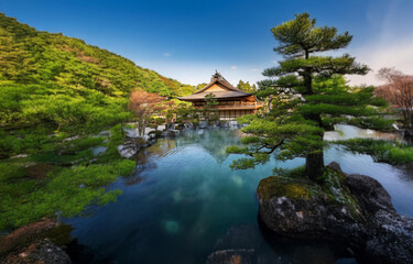 A tranquil pond surrounded by lush greenery and a wooden structure, with a clear blue sky above.