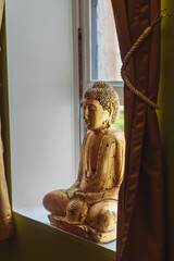 A golden Buddha statue sits in a meditative pose on a windowsill, framed by draped curtains,...