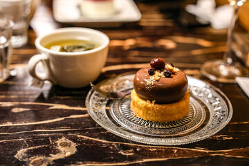 Chocolate Mousse Cake with Tea on Rustic Table. A gourmet chocolate mousse cake topped with glazed berries and nuts, elegantly served on a glass plate, accompanied by a cup of hot tea.