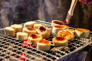 Grilling Tofu with Sauce Brush. A close-up shot of tofu rounds grilling over charcoal. The tofu pieces are being glazed with a brush, adding a layer of sauce that will caramelize beautifully.