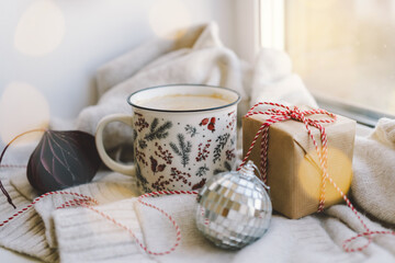 A warm beverage sits in a decorative mug next to a gift and holiday ornaments, creating a cozy atmosphere perfect for winter relaxation.