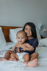A little girl with a joyful expression is gently holding a baby on a cozy bed, creating a heartwarming scene filled with love