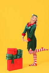 Happy young woman dressed as elf with Christmas gifts on yellow background
