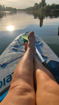 Bare legs of woman on SUP board on surface of river or lake with sun reflecting on warm summer evening. Person swimming on SUP board on pond on calm day. Tourism travel rest sport lifestyle Vertical