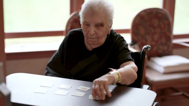 An elderly woman with white hair sits in a wheelchair, her hand reaching for a card on a table. The table has a few other cards scattered on it, and the woman is wearing a black shirt with a sling aro