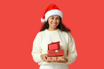 Happy African-American woman in Santa hat and with Christmas gifts on red background