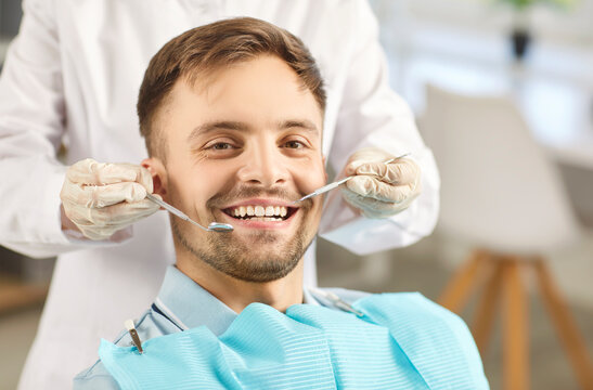 Portrait of male patient looking cheerful at camera in modern dentistry clinic during medical check up in medical center. Dentist examining young man teeth with dental tools. Dental health care.