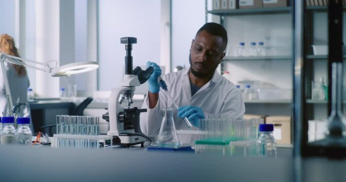 Medical science lab: African American microbiologist takes liquid from test tube, looks under microscope, does analysis of sample using high-tech equipment. Female scientist works in the background.