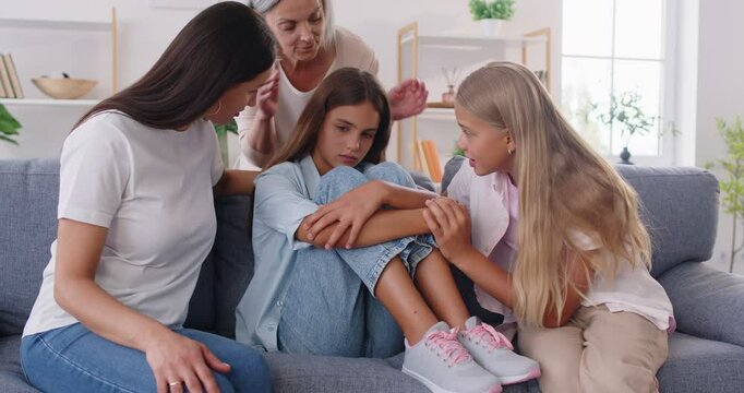 Anxious family gathering around sad teenage girl on sofa asking about her problems. Sister mother and grandmother comforting her in difficult life situation with tender words, stroking and supporting.