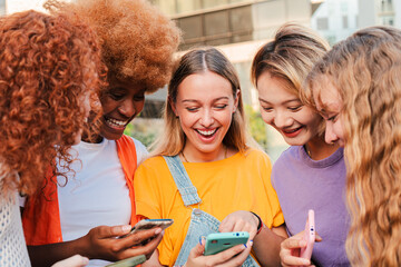 Group of diverse young women happily using their smartphones together outdoors, sharing and...