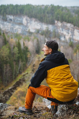 A man wearing a bright yellow and black jacket is sitting comfortably on a large rock while enjoying the stunning view of a vast, sprawling forest that stretches out below him