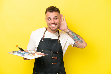 Young artist caucasian man holding a palette isolated on yellow background listening to something...