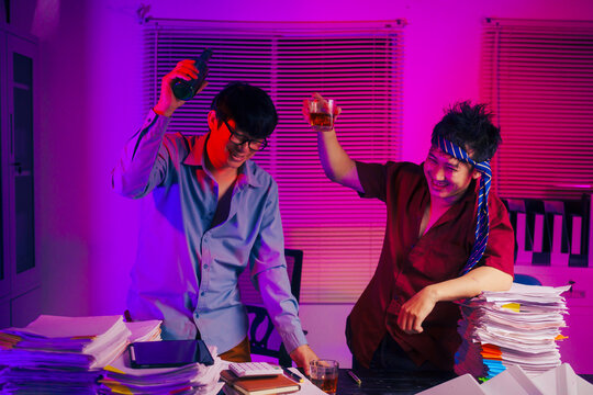 Two men are working late at the office, drinking beer while checking documents. They are overwhelmed with a lot of work, tired, and stressed as they continue to work through the night.