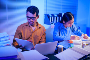 A businessman and woman are working overtime at their desks in  office at night.They are busy, stressed, overloaded with too much work, checking documents and working on laptops while feeling tired