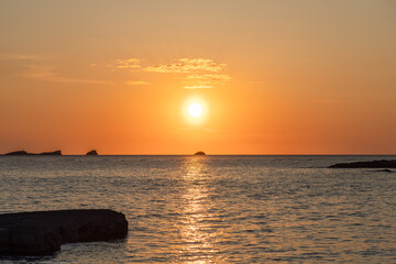 A captivating sunset at Cala Conta beach in Ibiza, Spain, features the sun setting over calm waters with a golden glow. Silhouetted islands dot the horizon, and the tranquil sea