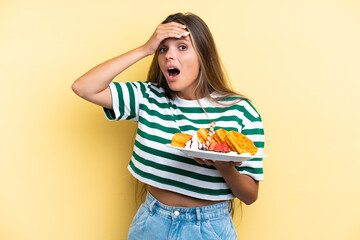 Young caucasian woman holding waffles isolated on yellow background doing surprise gesture while looking to the side