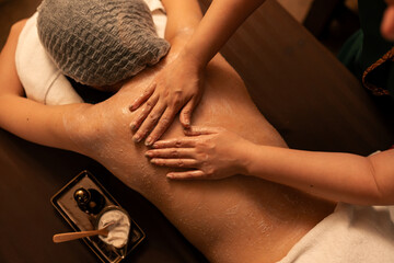 Top view, Motion blur hand of masseuse applying cream salt scrub on customer back at a cosmetology spa center. Woman customer enjoy soothing skincare scrub massage in serene ambiance of spa salon.