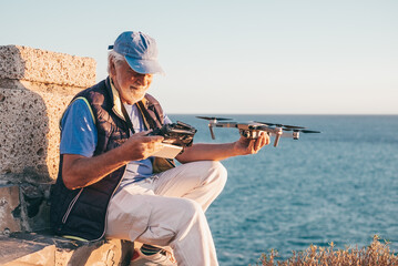 Senior man outdoors at sunset holding a drone and the remote controller in hands. Aerial photographs. Sea on background, horizon over water © luciano