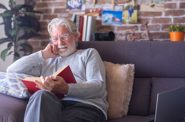 Smiling senior bearded man with eyeglasses relaxing on sofa at home reading a book. Retirement lifestyle concept