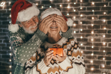 Lovely old man surprises his wife with a gift, seniors at home enjoying Christmas together in December wearing Santa hats. Elderly man and woman celebrate holidays