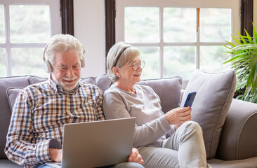 Smiling white-haired 70-year-old couple at home using technological devices, man uses laptop watching video, woman using phone. Older people use new technologies