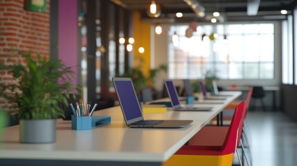 Bright and colorful shared workspace featuring a row of laptops, vibrant chairs, and office plants in a contemporary setting.