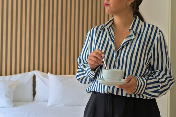  Woman stirring coffee in a modern hotel room with striped shirt and white bed linens