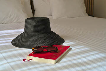 A black sunhat, sunglasses, and a travel journal on a bed with a bottle of champagne in the background, suggesting a luxury vacation.