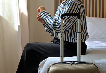 Woman sitting on hotel bed adjusting curtain with suitcase, preparing for travel