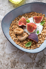 Bowl of granola served with fresh and dried fig fruits and greek yogurt, vertical shot on a light-brown granite surface, middle closeup