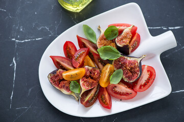 Salad with heirloom tomatoes, fresh figs and basil on a shovel-shaped plate, horizontal shot on a black marble background, elevated view