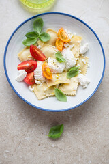 Blue and white plate with ravioli, ricotta, tomatoes and fresh basil, vertical shot on a beige stone background, high angle view