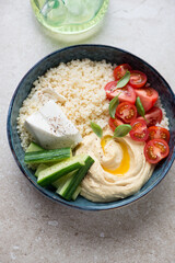 Mediterranean hummus bowl with couscous, feta and fresh vegetables, vertical shot on a beige stone background, elevated view