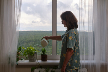 A woman waters flowers from a watering can on the windowsill near a large window