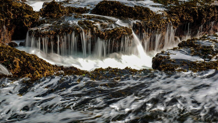 The crashing waves at Karang Bokor Sawarna Beach flowing over the coral rocks become a mini waterfall. The rough texture of the coral rocks and green moss add dimension to the scenery.