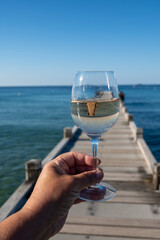 Hand with glass of cold rose wine from Provence and wooden yacht boota pier on white sandy beach Plage de Pampelonne near Saint-Tropez, summer vacation in France