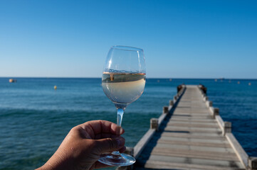 Hand with glass of cold rose wine from Provence and wooden yacht boota pier on white sandy beach Plage de Pampelonne near Saint-Tropez, summer vacation in France
