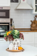 Still-life. A bouquet of decorative cabbage in a vase, pumpkins and white houses on the table in the interior of a home kitchen. A cozy autumn concept.